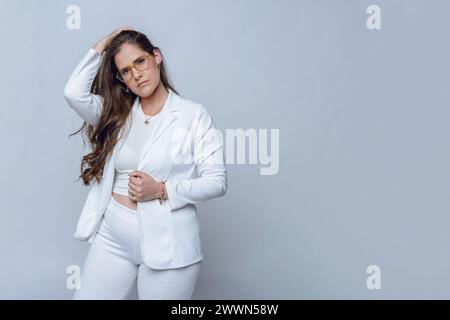 Attraktive junge lateinische Geschäftsfrau mit langen Haaren und Brille, posiert in weißen, eleganten Kleidern, mit einer Hand auf dem Kopf und blickt auf Kameras Stockfoto