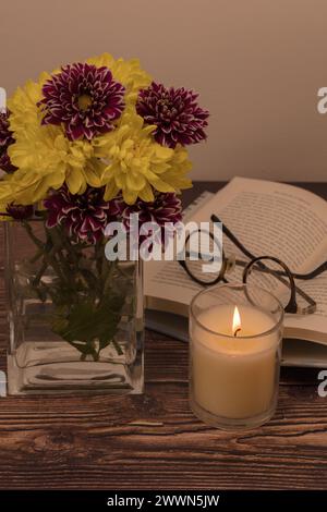 Foto im Portraitformat/Bild einer Brille auf einem offenen gebundenen Buch mit beleuchteter Kerze und Chrysanthemen in einer quadratischen Vase auf einem Vintage-Holztisch Stockfoto