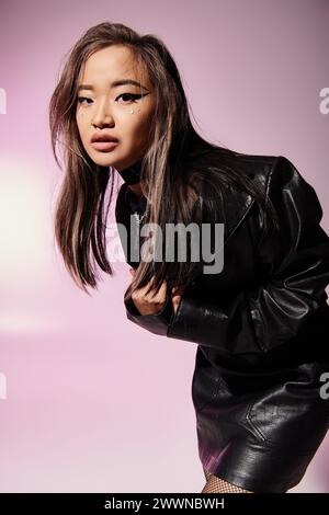 Hübsche Frau in schwarzem Leder-Outfit mit schwerem Make-up seitlich nach vorne gelehnt auf lilafarbenem Hintergrund Stockfoto
