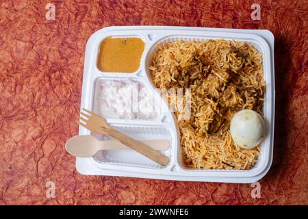 Indisches Hühnchen Biryani verpackt in recycelter Box mit Raita und Soße und Holzlöffel für die Lieferung nach Hause. Stockfoto
