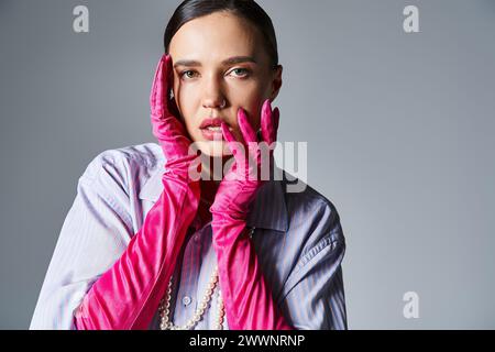 Träumende Frau in violettem Hemd und Perlen, rosa trendigen Handschuhen, die ihre Lippen berührt und auf Grau steht Stockfoto
