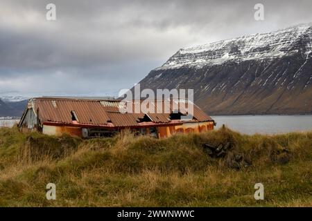 Verlassenes Haus mit einem rostigen und kaputten Dach. Herbstgras herum. Gelegen an der Küste eines Fjords mit einem großen Berg auf der anderen Küste, mit Schnee o Stockfoto