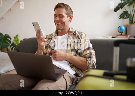 Ein lächelnder, gutaussehender Mann, der in Urlaub geht, einen Anruf macht, einen Laptop hält, einen Koffer für eine Reise ins Ausland vorbereitet Stockfoto