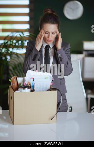 Neuer Job. Besorgte, moderne Angestellte in einem modernen grünen Büro in grauem Business-Anzug mit persönlichen Sachen im Karton. Stockfoto