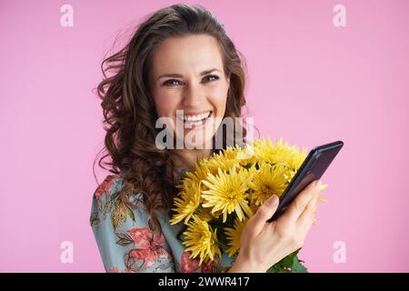 Porträt der glücklichen stilvollen Frau im Blumenkleid mit gelben Chrysanthemen Blumen mit Smartphone-App isoliert auf rosa Hintergrund. Stockfoto