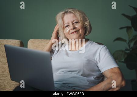 Lächelnde ältere Frau, die im Wohnzimmer auf einem Sessel sitzt. Glückliche, selbstbewusste Seniorin, die zu Hause am pc arbeitet und auf der Tastatur schreibt. Stockfoto