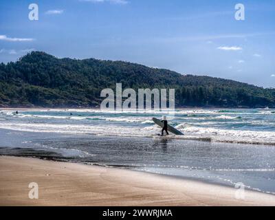 Ein Surfer läuft am Long Beach am Ufer des Wassers entlang, trägt ein Surfbrett unter einem Arm und blickt in die Ferne. Stockfoto