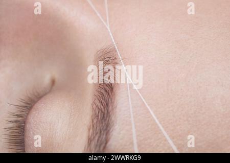 Die Make-up-Künstlerin zupft ihre Augenbrauen mit einer Fadennaht. Schönheitsbehandlungen für Gesichtspflege im Schönheitssalon. Stockfoto
