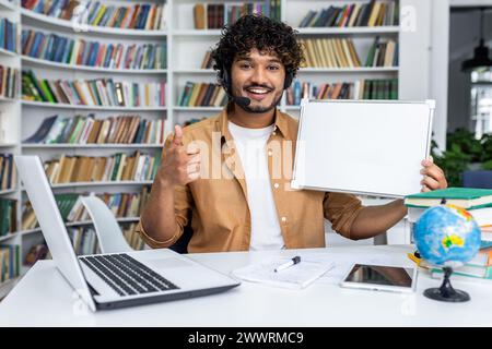 Lächelnder indischer Mann mit schnurlosem Headset, der ein leeres Whiteboard hält und die Daumen hoch vor einem modernen Laptop mit verschiedenen Gegenständen auf dem Schreibtisch hält. Hintergrund von Bücherregalen und Home Office. Stockfoto