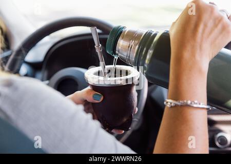 Frau, die in einem Auto sitzt und Yerba Mate Wasser aus einer Thermoskanne in einen Mate Becher gießt. Stockfoto