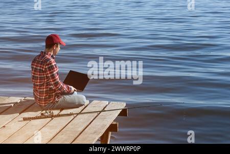 Ein Fischer arbeitet an einem Laptop, sitzt auf einem hölzernen Pier in der Nähe des Sees, daneben befindet sich eine Angelstange. Stockfoto