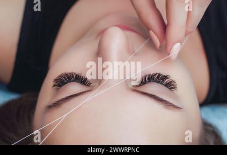 Die Make-up-Künstlerin zupft ihre Augenbrauen mit einer Fadennaht. Schönheitsbehandlungen für Gesichtspflege im Schönheitssalon. Stockfoto