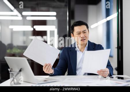 Ein knackiger Mann, der Dokumente hält, während er am weißen Schreibtisch im Büro sitzt. Gutaussehender asiatischer Mann, der auf dem Hintergrund auf der Glaswand des Business Centers arbeitet. Konzept von Technologie und Mensch. Stockfoto