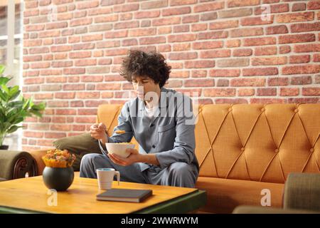 Junger Mann im Pyjama, der Cornflakes zum Frühstück isst und auf einem Sofa sitzt Stockfoto