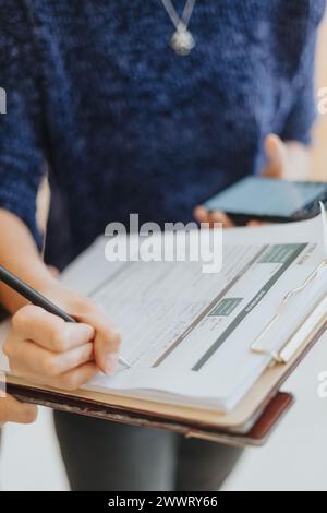 Die weibliche Mitarbeiterin prüft die Papiere genau, während sie ein Smartphone hält, und zeigt Effizienz und Multitasking in einer modernen Büroumgebung. Stockfoto