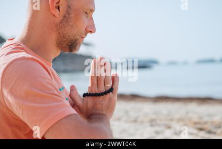Ruhiger Mann, der mit Namaste-Händen zusammen sitzt und tiefe Atemübungen macht und in den frühen Morgenstunden am Sandstrand mit ruhigen Meereswellen meditiert. Stockfoto