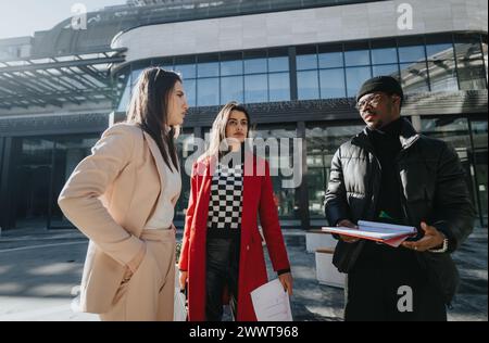 Draußen wird ein multikulturelles Geschäftsteam gezeigt, mit zwei lächelnden Frauen und einem fokussierten Mann, der Notizen macht. Stockfoto