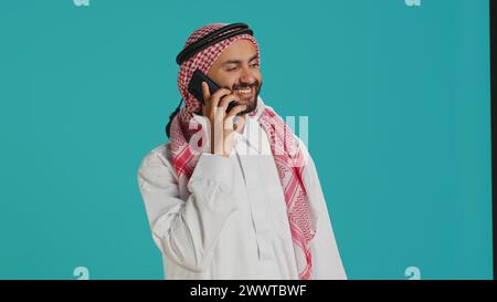 Muslimischer Mann in traditioneller Kleidung, der auf dem Handy schreibt, während er stolz im Studio posiert. Erwachsener aus dem Nahen Osten, gekleidet mit einem karierten Kopftuch, das online Nachrichten per Telefon sendet. Stockfoto