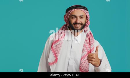 Ein selbstbewusster Mann zeigt sich wie ein Schild im Studio, gibt Daumen hoch und stimmt überein, während er die traditionelle arabische Kultur repräsentiert. Ein positiver Mann aus dem Nahen Osten trägt islamische Kostüme und Kopftuch. Stockfoto
