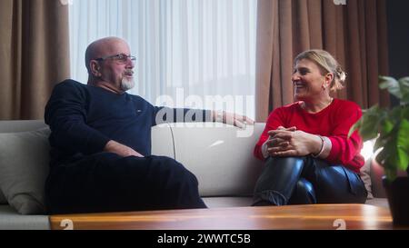 Ein älteres Familienpaar sitzt zu Hause auf der Couch, redet und spricht über den Ruhestand. Reife Frau hört dem sprechenden Ehemann zu, lächelt und genießt Beziehungen Stockfoto