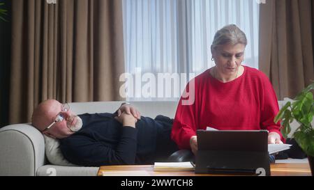 Eine Frau mittleren Alters sitzt zu Hause auf dem Sofa und überprüft Informationen auf Dokumenten, macht Notizen, macht Papiere. Lächelnde alte Frau reden und arbeiten mit Ehemann, dem ich Stockfoto