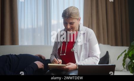 Professionelle Ärztin mittleren Alters im weißen Mantel mit Stethoskop, die ältere Patienten zu Hause untersucht, Medikamente verschreibt Stockfoto