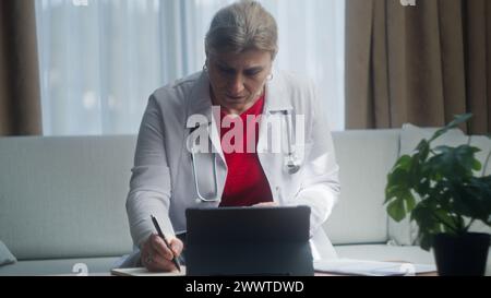 Leitende Ärztin im weißen Mantel Arbeit am Arbeitsplatz auf dem Sofa sitzen, Notizen schreiben, Bericht erstellen, Patientenkarte ausfüllen, Medikamente verschreiben Stockfoto