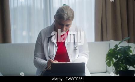 Reife Ärztin im weißen Mantel Arbeit am Arbeitsplatz auf dem Sofa sitzen, Notizen schreiben, Bericht erstellen, Patientenkarte ausfüllen, Medikamente verschreiben Stockfoto