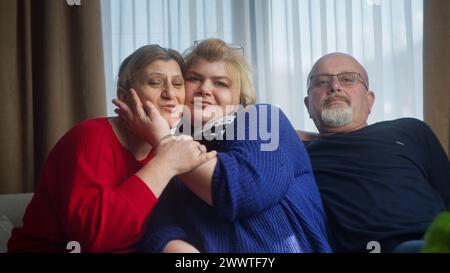 Glückliche Familie, die auf dem Sofa sitzt, lächelnd in die Kamera schaut, posiert, Spaß hat, lustige Gesten macht. Glückliches Familienporträt Stockfoto