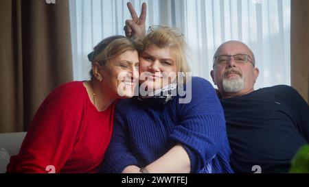 Fröhliche, glückliche Familie, die auf dem Sofa sitzt, lächelnd in die Kamera schaut, posiert, Spaß hat, lustige Gesten macht. Glückliches Familienporträt Stockfoto