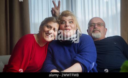 Überglücklich glückte Familie, die auf dem Sofa sitzt und lächelnd in die Kamera blickt, posiert, Spaß hat, lustige Gesten macht. Glückliches Familienporträt Stockfoto