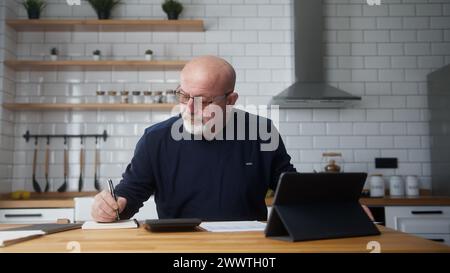 Ein alter Mann sitzt in der Küche mit einem Tablet-Gerät, studiert Finanzunterlagen mit einem Rechner, zählt Ausgaben, führt Budgetkontrolle durch, verwaltet Finanzen, Bankgeschäfte Stockfoto