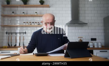 Älterer Mann sitzt mit einem Tablet in der Küche, studiert Finanzunterlagen mit einem Rechner, zählt Ausgaben, führt Budgetkontrolle durch, verwaltet Finanzen, Bank Stockfoto