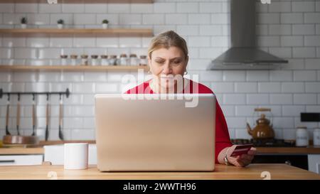 Ältere Frau, die in der Küche sitzt, geben Sie die Kreditkartennummer auf dem Laptop ein, um online einzukaufen, macht sichere, einfache elektronische Bezahlung in entfernter Entfernung Stockfoto