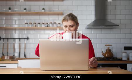 Reife Frau sitzt in der Küche mit Laptop, verbringt Freizeit im Internet, genießt Kommunikation in sozialen Medien, ruht zu Hause aus. Kabellos, modern Stockfoto