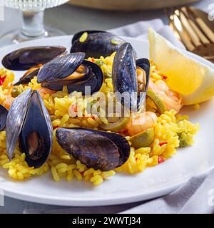 Paella mit Muscheln und Garnelen, auf einem weißen Teller, serviert mit Zitrone und Weißwein, quadratisch, Nahaufnahme Stockfoto