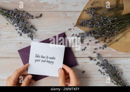 NEHMEN SIE SICH DIE ZEIT, UM IHRE SEELE GLÜCKLICH ZU MACHEN, indem Sie auf einer unterstützenden Nachricht auf Papier Notizen aus dem grünen Umschlag erinnern. Flache Zusammensetzung trockene Lavendelblüten. Konzept von innerem Glück, Verlangsamung digitaler Detox persönliche Erfüllung. Draufsicht Stockfoto