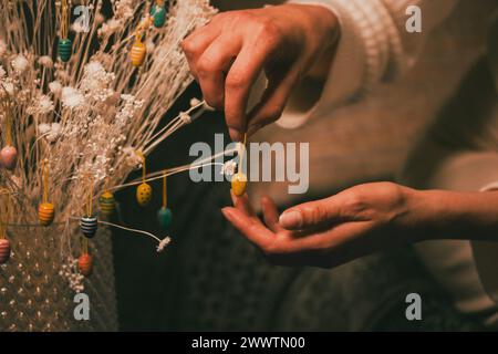 Eine Frau hängt ein gelbes dekoratives Osterei an einem Strauß trockener weißer Blumen im Inneren. Festliche Einrichtung. Die Hand der Frau hält einen Spielzeuganhänger. Stockfoto