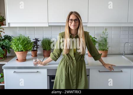 Zufriedene Frau mittleren Alters in Brille mit einem Glas Wasser, die in die in der Küche stehende Kamera blickt. Stockfoto