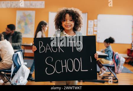 Stolzer Schüler, der am ersten Tag der Grundschule vor dem Klassenzimmer stand und ein Schild mit dem Rücken zur Schule hielt. Junge freut sich, eine neue Klasse zu beginnen Stockfoto