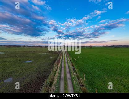 Eine bezaubernde Landstraße erstreckt sich in die Ferne, flankiert von lebhaften grünen Feldern unter einem weiten Himmel dramatischer Wolken. Ländliche Straße führt durch grüne Felder unter einem dynamischen bewölkten Himmel. Hochwertige Fotos Stockfoto