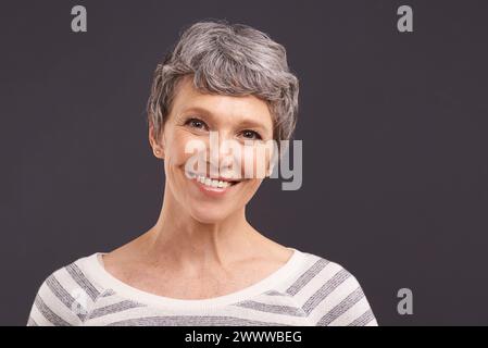 Porträt, Lächeln und ältere Frau mit Ruhestand, Glück und lässigem Outfit auf grauem Studiohintergrund. Gesicht, Rentner oder ältere Person mit Mockup Stockfoto