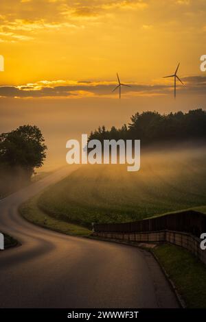 Eine Straße, die in den Nebel führt, und ein schöner Sonnenaufgang mit Windturbinen im Hintergrund (Bayern, Deutschland) mit Kopierraum Stockfoto