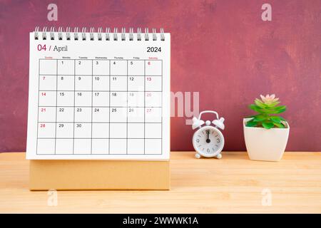Monatlicher Schreibtischkalender April für 2024 Jahre und Wecker auf Holztisch mit rotem Hintergrund. Stockfoto