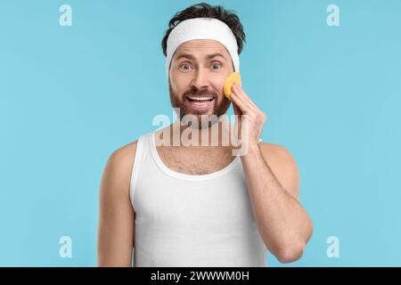 Mann mit Stirnband, der sein Gesicht mit Schwamm auf hellblauem Hintergrund wäscht Stockfoto