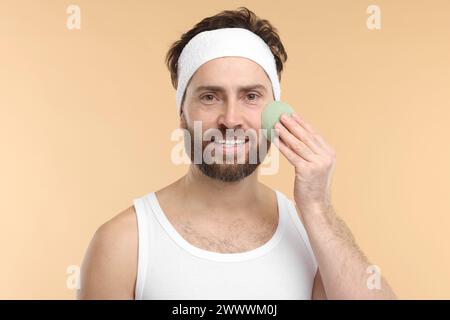 Mann mit Stirnband, der sein Gesicht mit einem Schwamm auf beigem Hintergrund wäscht Stockfoto
