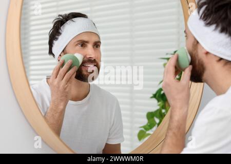 Mann mit Stirnband, der sein Gesicht mit einem Schwamm in der Nähe des Spiegels im Badezimmer wäscht Stockfoto