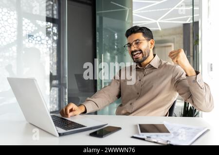 Erfolgreicher Geschäftsmann im Büro feiert Sieg und Triumph, Mann, der glückliche Nachrichten von Laptop liest, Unternehmer, der am Arbeitsplatz arbeitet, zufrieden mit Leistungsergebnissen. Stockfoto