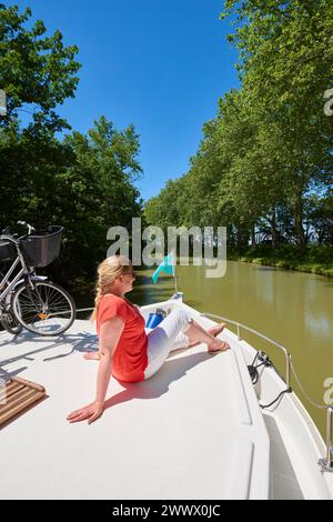 Binnenschifffahrt auf dem Canal du Midi. Frau, die sich entspannt und die Sonne auf dem Vordeck genießt, neben ihrem Fahrrad. Der Canal du Midi ist als UNESCO Wor eingetragen Stockfoto