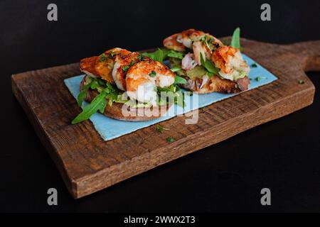 Gesunde Vorspeise oder Snack Garnelen Bruschetta mit Vollkornbrot auf Holzbrett auf schwarzem Hintergrund. Gebratene Garnelen und pürierte Avocado auf einem gerösteten Stockfoto